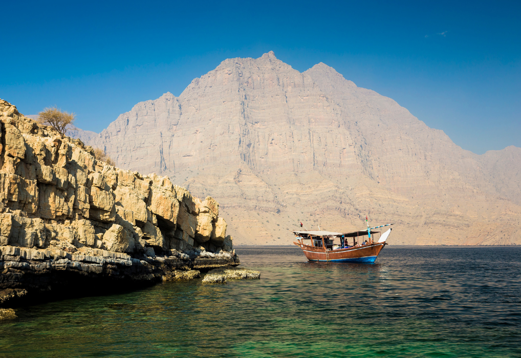 Traditional wooden boat sailing near rugged cliffs of Musandam Dibba with towering mountains in the background.