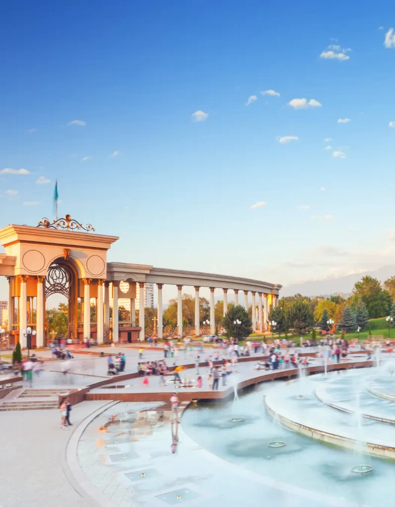 Panoramic view of Almaty's iconic park with fountains, bustling with locals and tourists, set against a clear blue sky.