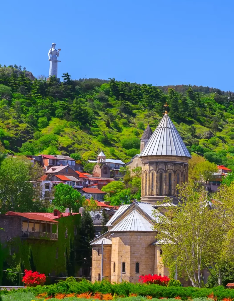 Panoramic view of Tbilisi, Georgia, featuring the Mother of Georgia statue and historic churches nestled among vibrant green hills.